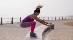 Vista laterale di una giovane donna caucasica che indossa vestiti sportivi saltando su un muro basso durante un allenamento in una giornata di sole in riva al mare — Foto stock