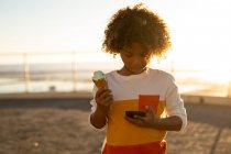 Frontansicht eines vorpubertären Jungen, der ein Eis in der Hand hält und auf ein Smartphone am Meer blickt, das von der untergehenden Sonne hinterleuchtet wird — Stockfoto
