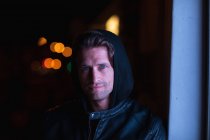 Portrait of a smiling young Caucasian man wearing a hoodie looking to camera leaning on a wall in an urban street at night — Stock Photo