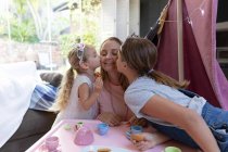 Front view of a young Caucasian woman with her tween and younger daughters having a dolls tea party at home and kissing their mother — Stock Photo