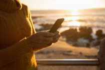 Вид збоку крупним планом жінка використовує смартфон біля моря на заході сонця — стокове фото