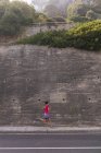 Vista lateral de uma jovem caucasiana vestindo roupas esportivas passando por uma parede de retenção alta durante um treino em um dia ensolarado — Fotografia de Stock