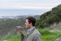 Vista lateral de cerca de un joven caucásico disfrutando de un día en el campo junto al mar, hablando en su teléfono inteligente - foto de stock