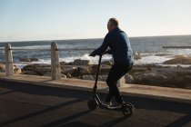 Vista trasera de un hombre caucásico maduro montando un e scooter junto al mar al atardecer - foto de stock
