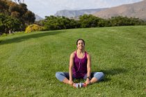 Vista frontale di una giovane donna caucasica che indossa abiti sportivi seduta sull'erba, tenendo i piedi, allungando e ascoltando musica mentre si allena in un parco — Foto stock