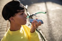 Вид збоку на модний молодий змішаний гонка транссексуал дорослий на вулиці, розмовляючи на смартфоні в береті з велосипедом на задньому плані — стокове фото