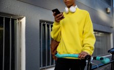 Vista frontal de un joven transexual de raza mixta de moda en la calle, enviando mensajes de texto en el teléfono inteligente y sosteniendo una bicicleta - foto de stock