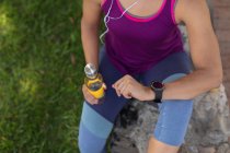 Піднятий вигляд середньої секції жінки в спортивному одязі, що тримає пляшку води, перевіряє її смарт-годинник і слухає музику на навушниках, працюючи сонячно в парку — стокове фото