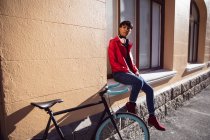 Vista frontal de um jovem adulto transexual de raça mista na moda na rua, sentado em uma soleira da janela ao lado de uma bicicleta — Fotografia de Stock