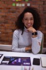 Портрет крупным планом молодой женщины смешанной расы, сидящей за столом, опирающейся на руку и улыбающейся в камеру, работающей в офисе творческого бизнеса — стоковое фото