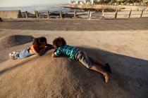 Vue latérale d'une jeune femme métissée et de son fils pré-adolescent jouissant de temps ensemble jouant sur une aire de jeux au bord de la mer, couchés sur le sol et se regardant par une journée ensoleillée — Photo de stock