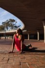 Nahaufnahme einer lächelnden jungen kaukasischen Frau in Sportkleidung, die Musik über Kopfhörer hört, sich auf ihre Hände stützt und ihren Rücken während eines Trainings an einem sonnigen Tag in einem Park streckt — Stockfoto
