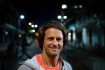 Portrait of a young Caucasian man wearing earphones smiling to camera in the street during his late evening workout — Stock Photo