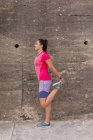 Вид збоку молоді кавказька жінка носить спортивний одяг стоячи перед стіною на вулиці, тримаючи ногу і розтягуючи ногу під час тренування — стокове фото