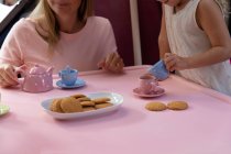 Vista frontal de una joven mujer caucásica con su hija pequeña teniendo una fiesta de té de muñecas en casa - foto de stock