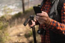 Vista lateral sección media del hombre usando un teléfono inteligente y sosteniendo bastones nórdicos en un entorno rural - foto de stock