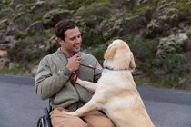Vista frontal de cerca de un joven caucásico en silla de ruedas dando un paseo con su perro en el campo, sonriendo al perro, que está de pie sobre sus patas traseras - foto de stock