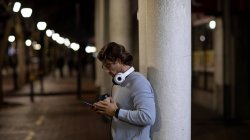 Vista lateral de um jovem caucasiano de pé em uma rua à noite com fones de ouvido, usando um smartphone e segurando um café takeaway — Fotografia de Stock