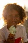 Ritratto di un adolescente sorridente con i capelli ricci che mangia un gelato in riva al mare, retroilluminato dal sole al tramonto — Foto stock