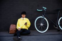Vista frontal de un joven transexual de raza mixta de moda en la calle, enviando mensajes de texto en el teléfono inteligente sentado junto a una bicicleta - foto de stock