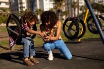 Вид спереди на молодую женщину смешанной расы и ее сына-подростка, наслаждающуюся временем вместе, играя на детской площадке, используя смартфон на качелях в солнечный день — стоковое фото