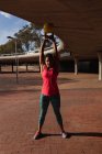 Вид спереди на молодую белую женщину в спортивной одежде, держащую гири над головой во время тренировки в солнечный день в парке — стоковое фото