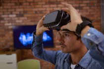Side view close up of a young Caucasian man sitting at a desk putting on a VR headset in the office of a creative business — Stock Photo