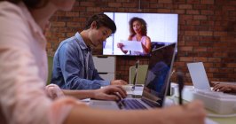 Side view close up of a young Caucasian woman and man working in the office of a creative business. A female colleague is visible on a wall mounted screen, communicating by video link — Stock Photo