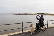 Rückansicht eines jungen kaukasischen Mannes im Rollstuhl, der mit seinem Hund am Meer spazieren geht und ein Foto macht — Stockfoto