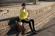 Nahaufnahme eines modischen jungen Transgender-Erwachsenen mit gemischter Rasse auf der Straße, der einen Laptop benutzt und mit dem Smartphone spricht — Stockfoto