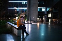 Вид збоку молодого Кавказького чоловіка, що сидить на стіні на вулиці будинку вночі слухати музику з навушниками на, дивлячись на смартфон — стокове фото