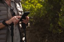 Вид сбоку средней части женщины, использующей смартфон и держащей палки для скандинавской ходьбы в сельской местности — стоковое фото