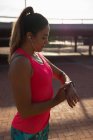 Vista lateral de cerca de una joven mujer caucásica sonriente que usa ropa deportiva revisando su reloj inteligente y escuchando música en los auriculares mientras hace ejercicio en un día soleado en un parque - foto de stock