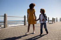 Вид спереди на молодую расовую женщину и ее сына-подростка, наслаждающихся морем, держащихся за руки и идущих в солнечный день — стоковое фото