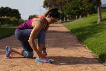 Seitenansicht einer jungen kaukasischen Frau in Sportkleidung, die kniet und ihren Schuh bindet, während sie Musik über Kopfhörer hört, bevor sie an einem sonnigen Tag in einem Park trainiert — Stockfoto