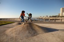 Vista lateral de una joven mestiza y su hijo preadolescente disfrutando del tiempo juntos jugando en un parque infantil junto al mar, el niño saltando en un día soleado - foto de stock