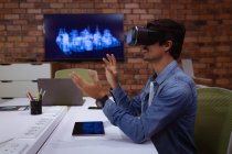 Side view close up of a young Caucasian man sitting at a desk wearing a VR headset with his hands raised, in the office of a creative business — Stock Photo