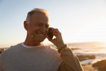 Vista frontal de cerca de un hombre caucásico maduro hablando por teléfono junto al mar al atardecer - foto de stock