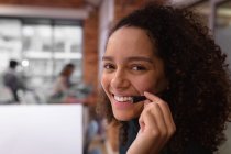 Ritratto ravvicinato di una giovane donna di razza mista sorridente che lavora nell'ufficio di un'azienda creativa con un auricolare per il telefono e che guarda alla telecamera, con colleghi che lavorano sullo sfondo — Foto stock