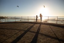 Rear view of a mixed race woman and her pre-teen son enjoying time together by the sea, admiring the view from a promenade on a sunny day, backlit by the setting sun — Stock Photo