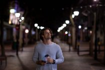 Вид на молоду Кавказьку людину, що ходить на вулиці вночі, тримаючи смартфон і каву на винос і оглядаючись — стокове фото