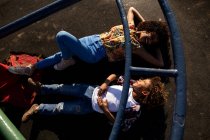 Передній вигляд молодої змішаної раси жінки та її попереднього сина насолоджуються разом грою на ігровому майданчику, лежачи на землі під скелелазінням на сонячний день — стокове фото