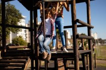 Vista frontale di una giovane donna di razza mista e del suo figlio pre-adolescente che si divertono insieme giocando in un parco giochi su una cornice di arrampicata in una giornata di sole — Foto stock