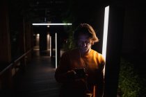 Frontansicht eines jungen kaukasischen Mannes, der nachts auf einer Straße steht, Musik mit Kopfhörern hört und auf ein Smartphone blickt — Stockfoto
