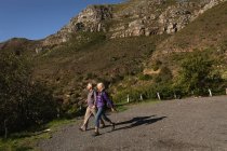 Side view of a mature Caucasian man and woman holding hands and walking in a rural setting, with mountains and a blue sky behind them — Stock Photo