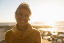 Porträt einer reifen kaukasischen Frau, die bei Sonnenuntergang am Meer in die Kamera lächelt — Stockfoto