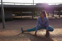 Nahaufnahme einer jungen kaukasischen Frau in Sportkleidung, die sich hinhockt und den Boden berührt, während sie in einem Park im Gegenlicht der Sonne trainiert — Stockfoto