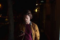 Frontansicht eines jungen kaukasischen Mannes, der nachts mit einem Smartphone auf einer Straße steht — Stockfoto