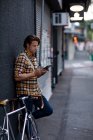 Seitenansicht eines jungen kaukasischen Mannes, der auf seinem abendlichen Heimweg einen Tablet-Computer mit einem Fahrrad neben sich hält und sich an eine Wand in einer städtischen Straße lehnt — Stockfoto