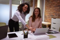 Front view close up of a young mixed race woman standing and a young Caucasian woman sitting at a desk, working together at a laptop in the office of a creative business, both looking to camera smiling — Stock Photo